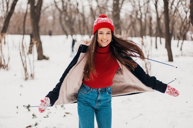 赤いミトンと冬のコートを着て、雪の中で公園を歩いて、暖かい服を着たニット帽の若いかわいい笑顔の幸せな女性