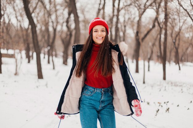 Young pretty smiling happy woman in red mittens and knitted hat wearing winter coat, walking in park in snow, warm clothes