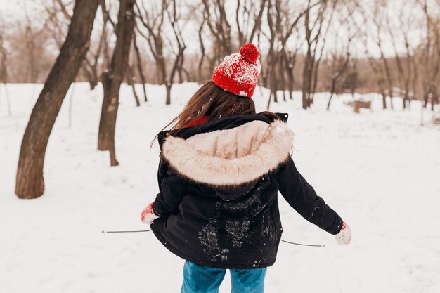 Молодая довольно улыбающаяся счастливая женщина в красных рукавицах и вязаной шапке в зимнем пальто, гуляет в парке в снегу, теплой одежде