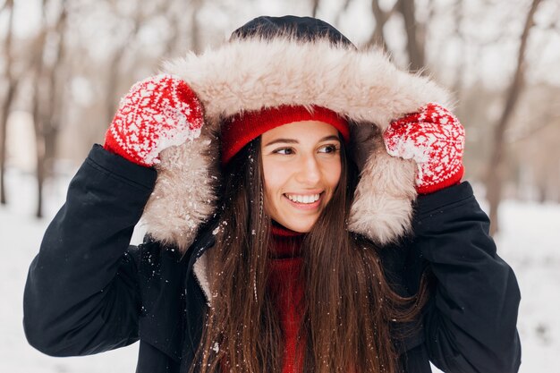 赤いミトンと毛皮のフード付きの冬のコートを着て、雪の中で公園を歩いて、暖かい服を着たニット帽の若いかわいい笑顔の幸せな女性