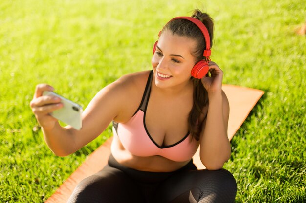 핑크색 스포티 상의와 레깅스에 빨간 헤드폰을 끼고 웃고 있는 젊고 예쁜 여성은 공원의 푸른 잔디에서 시간을 보내면서 행복하게 휴대폰으로 사진을 찍고 있습니다