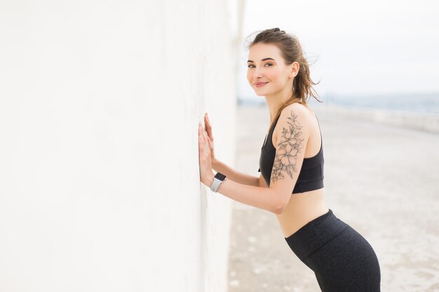 Young pretty smiling woman in dark gray sporty top and leggings joyfully looking in camera while leaning on wall doing exercises outdoor