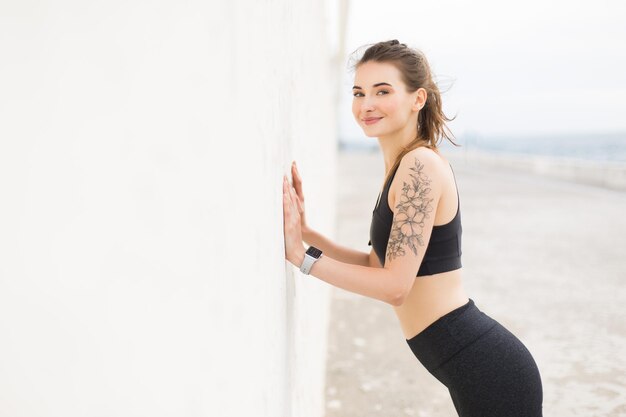 Молодая симпатичная улыбающаяся женщина в темно-сером спортивном топе и леггинсах радостно смотрит в камеру, опираясь на стену, делая упражнения на свежем воздухе