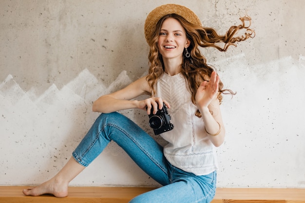 Молодая симпатичная улыбающаяся женщина в синих джинсовых джинсах и белой рубашке сидит у стены в соломенной шляпе