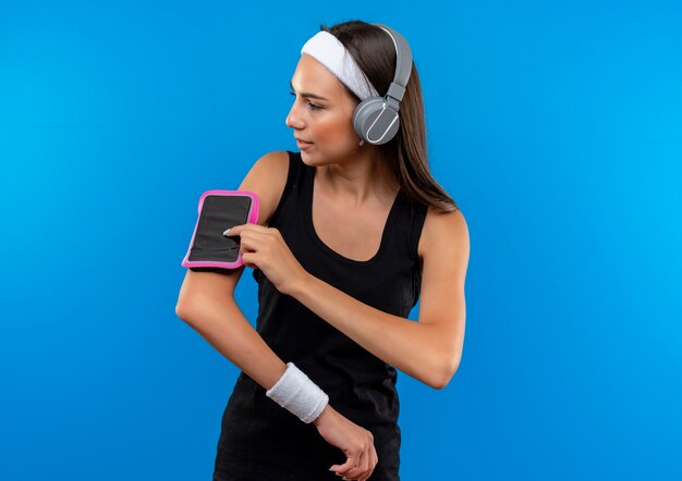 Young pretty sporty girl wearing headband and wristband and headphones touching phone armband looking at side 