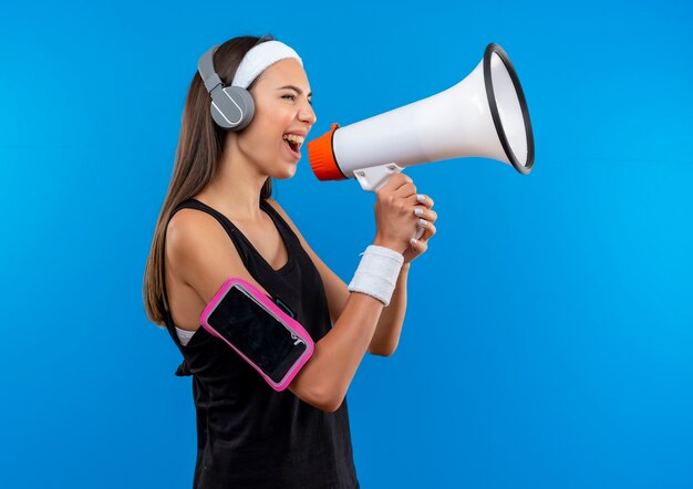 Young pretty sporty girl wearing headband and wristband and headphones with phone armband talking by speaker standing in profile view 
