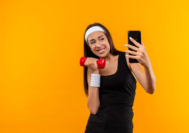 Young pretty sporty girl wearing headband and wristband holding dumbbell and taking selfie 