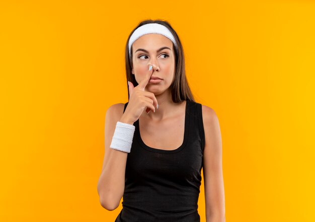Young pretty sporty girl wearing headband and wristband putting finger in nose and looking at side