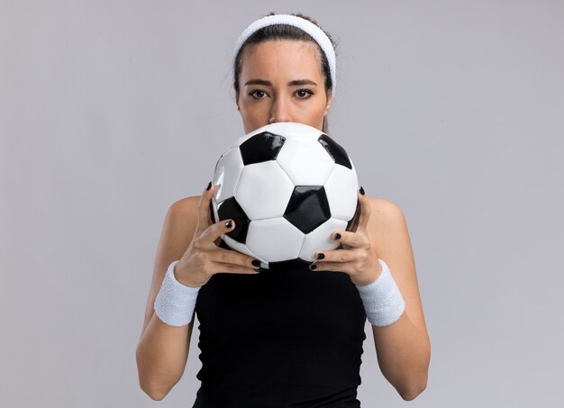 Young pretty sporty girl wearing headband and wristbands holding soccer ball from behind it 