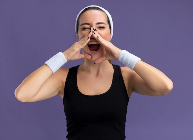 young pretty sporty girl wearing headband and wristbands keeping hands around mouth  calling out loud to someone isolated on purple wall