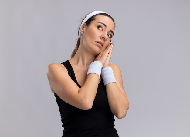 Young pretty sporty woman wearing headband and wristbands doing sleep gesture looking up isolated on white wall with copy space