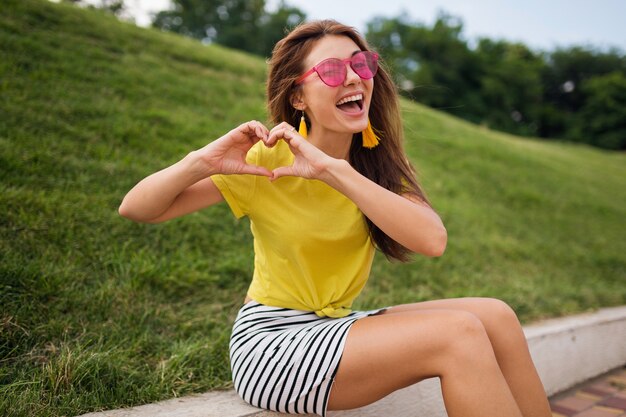 Молодая довольно стильная счастливая улыбающаяся женщина веселится в городском парке, позитивная, эмоциональная, в желтом топе, полосатой мини-юбке, розовых солнцезащитных очках, модная тенденция в летнем стиле, показывает знак сердца