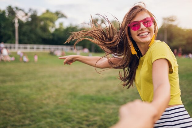 Young pretty stylish smiling woman having fun in city park, holding hand of boyfriend, follow me, positive, emotional, wearing yellow top, pink sunglasses, summer style fashion trend, positive emotion