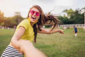 Free photo young pretty stylish smiling woman having fun in city park, holding hand of boyfriend, follow me, positive emotional, wearing yellow top, pink sunglasses, summer style fashion trend