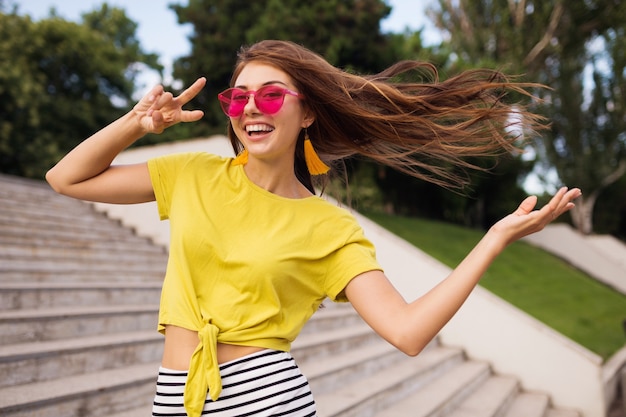 Young pretty stylish smiling woman having fun in city park, positive, emotional, wearing yellow top, striped mini skirt, pink sunglasses, summer style fashion trend, long hair, showing peace sign