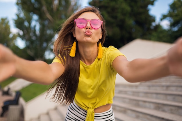 Young pretty stylish smiling woman making selfie in city park, positive, emotional, wearing yellow top, pink sunglasses, summer style fashion trend, long hair, having fun