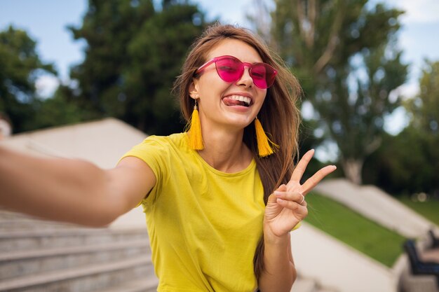 젊은 꽤 세련된 웃는 여자 도시 공원에서 셀카 만들기, 긍정적, 정서적, 노란색 탑, 핑크 선글라스, 여름 스타일 패션 트렌드, 긴 머리, 평화 기호 표시