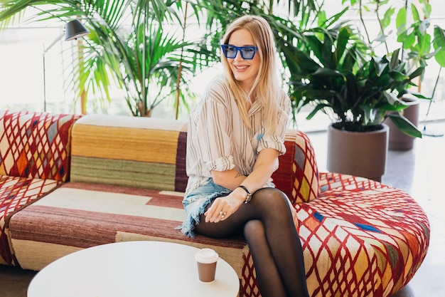 シャツと青いサングラスをかけているソファの上の流行に敏感なカフェに座っている若いかなりスタイリッシュな笑顔の女性