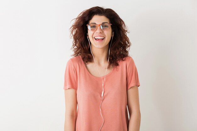 Young pretty stylish woman in glasses listening to music on earphones, curly hair, laughing, positive sincere emotion, happy, isolated, pink t-shirt