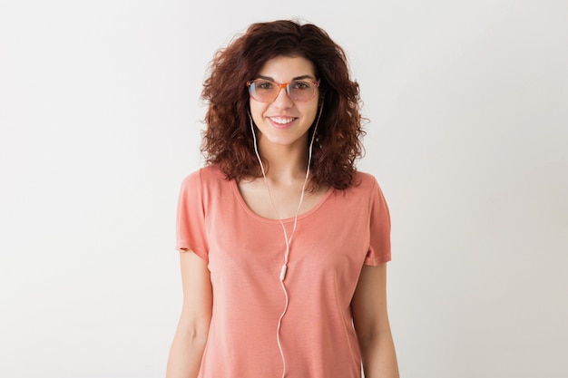 Young pretty stylish woman in glasses listening to music on earphones, curly hair, smiling, positive emotion, happy, isolated, pink t-shirt, motion