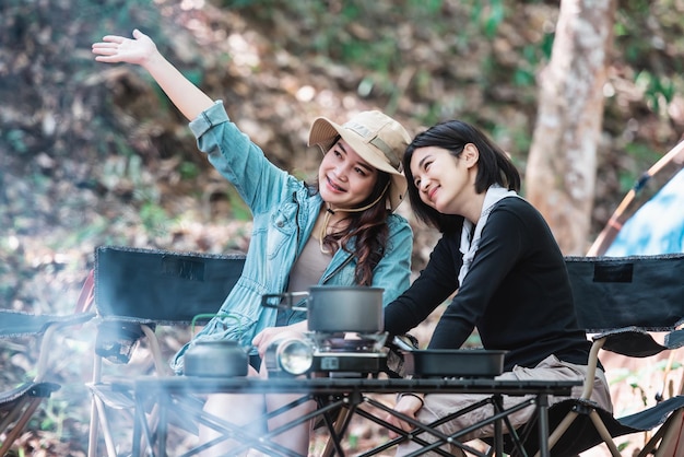 休日の休暇で森のキャンプテントに座っている間、カメラでかなり若い2人の女性の友人が写真を撮る若いアジアのグループの女性は屋外キャンプを旅行します