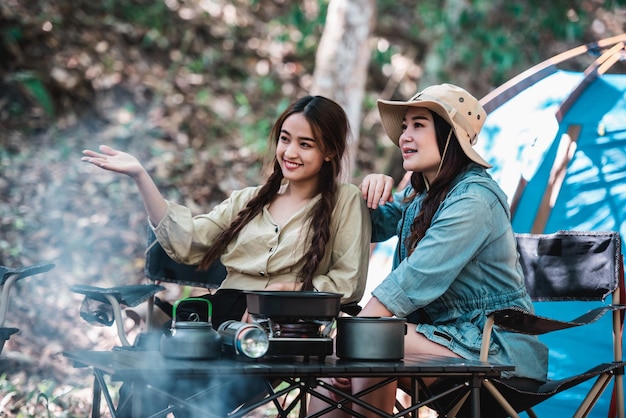 Young pretty taking photo by camera two female friends while sitting at the camping tent in forest on holiday vacation Young Asian group woman travel outdoor camping