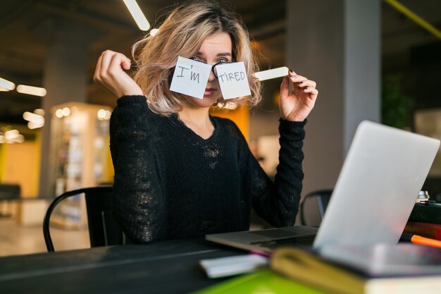 Young pretty tired woman with paper stickers on glasses sitting at table in black shirt working on laptop in co-working office, funny face emotion, problem, workplace, holding hands up