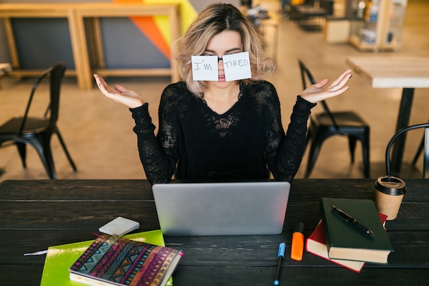 Young pretty tired woman with paper stickers on glasses sitting at table in black shirt working on laptop