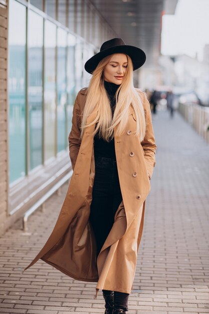Young pretty woman in black hat and beige coat walking by mall