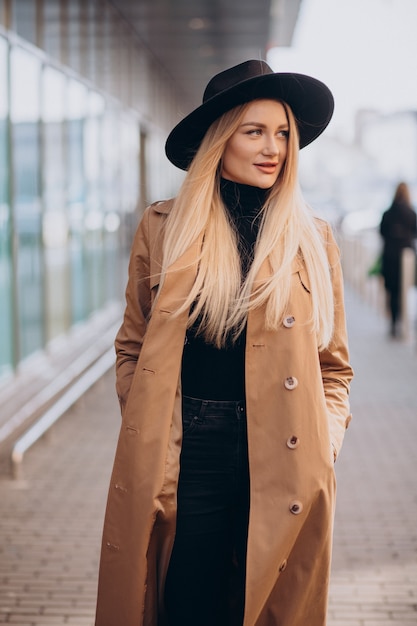 Young pretty woman in black hat and beige coat walking by mall