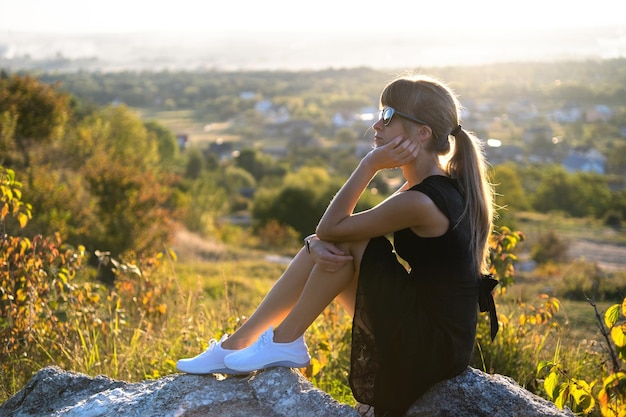 Young pretty woman in black short summer dress sitting on a rock relaxing outdoors at sunset. fashionable female enjoying warm evening in nature.