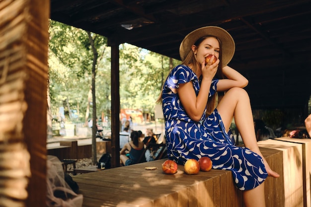 Young pretty woman in blue dress and hat barefoot eating peach joyfully looking in camera while sitting on wooden fence in city park