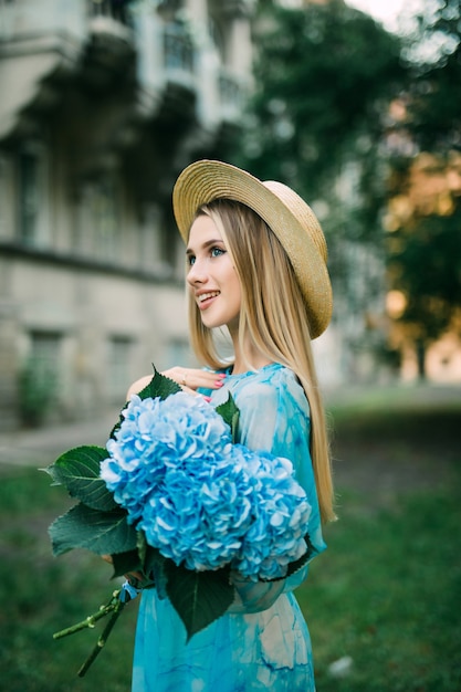 Giovane bella donna in abito blu che tiene i fiori di ortensia nella strada estiva