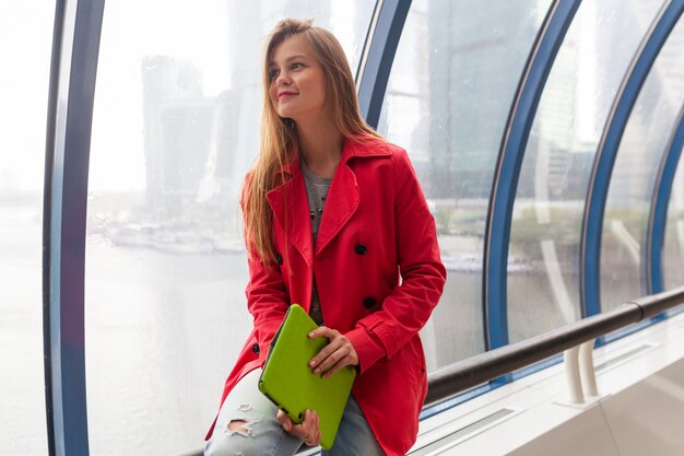 Young pretty woman in casual outfit holding tablet laptop in urban building, wearing jeans, pink trench jacket, sitting at window with city view