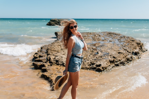 Giovane donna graziosa in vestito e cappello di paglia che cammina sulla spiaggia di onocean
