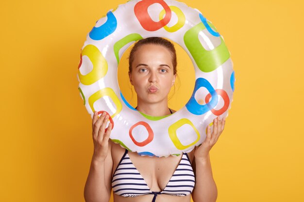 Young pretty woman feeling tired and frustrated, looking frustrated with problem, posing with pout lips, holding inflatable ring in front of face, stands against yellow wall.