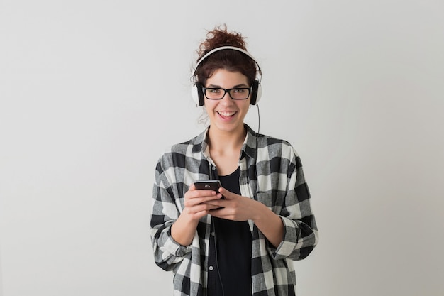 Young pretty woman in glasses, using smartphone, emotional, laughing, positive, happy, listening to music on headphones, isolated, checkered shirt, hipster style, student, looking in camera