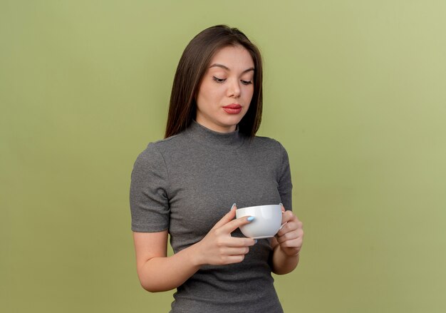 Free photo young pretty woman holding and looking at cup isolated on olive green with copy space