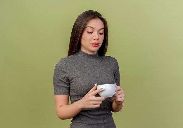young pretty woman holding and looking at cup isolated on olive green with copy space