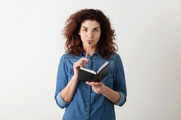 Young pretty woman holding notebook, pencil at lips, thinking, smiling, curly hair, pensive, happy, isolated