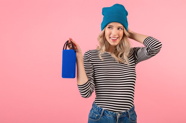 Young pretty woman holding wireless speaker listening to music wearing striped shirt and blue hat smiling happy positive mood posing on pink background 