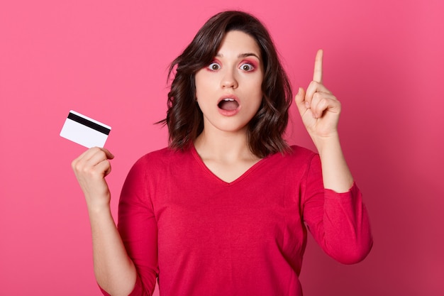 Young pretty woman looking astonished in disbelief, pointing up with her index finger, being unbelievable with credit card and widely opened mouth, female with surprised facial expression.