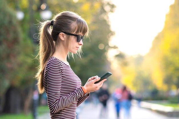 屋外の街の通りで暖かい秋の日に携帯電話でメッセージを送る若いきれいな女性。
