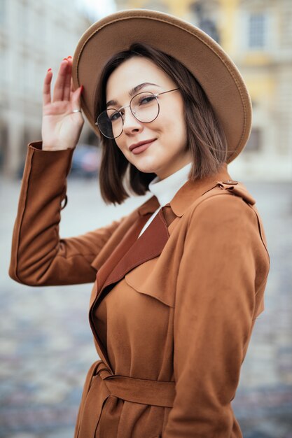 Young pretty woman in modern glasses and fashion hat and brown coat is posing in city centre