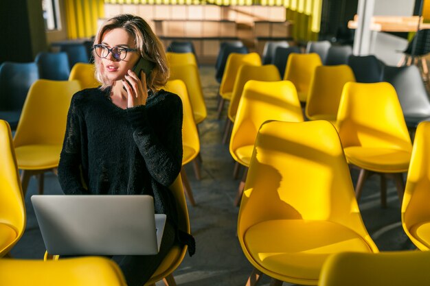 Young pretty woman sitting alone in co-working office, conference room, many yellow chairs, working at laptop, sunny, backlight, talking on phone, communication