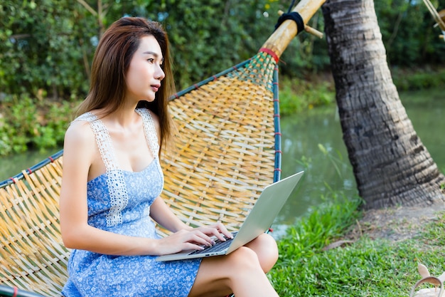 Молодая милая женщина сидя в бамбуковом гамаке используя компьтер-книжку