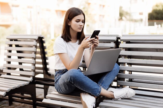 Young pretty woman sitting on bench in green park on summer day and reading text message on cell phone while using silver laptop.