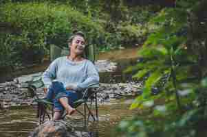 Free photo young pretty woman sitting on camping chair in stream and close her eyes for relaxtion she smile in nature forest while camping trip with happiness copy space