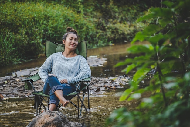 Young pretty woman sitting on camping chair in stream and close her eyes for relaxtion she smile in nature forest while camping trip with happiness copy space