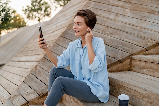 스마트폰을 사용하여 현대적인 공원 거리에서 노트북과 함께 앉아 있는 젊은 예쁜 여자